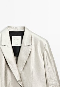Zilverkleurige, op maat gemaakte blazer met ingesneden kraag, een gladde textuur en een binnenvoering van zwart stof. Etiket zichtbaar bij de halslijn.