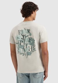 Beige t-shirt met korte mouwen, voorzien van een bloemenafbeelding in groen en blauw op de achterkant, met grote letters geïntegreerd in het ontwerp.