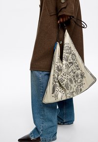 Kremowa torba w kształcie trójkąta z czarnymi liniami przedstawiającymi zwierzęta i kwiaty, z kontrastującą czarną krawędzią i dwoma teksturowanymi uchwytami do noszenia.