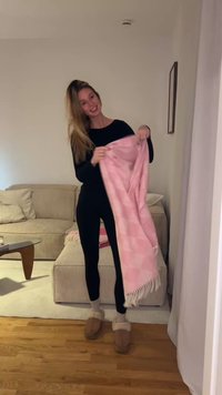 Ροζ κουβέρτα με σχέδια και φούντες που κρατιέται από ένα άτομο με μαύρα ρούχα. Το φόντο περιλαμβάνει έναν καναπέ με ρίγες και ξύλινο πάτωμα.