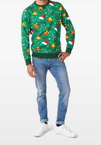 Suéter verde con patrones festivos que incluyen regalos, bastones de caramelo, campanas y gorros de Santa; puños y dobladillo acanalados, combinado con jeans azules y zapatos blancos.