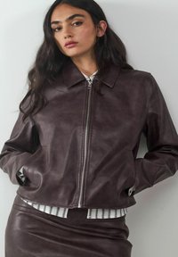 Bruine leren jack met een rits aan de voorkant, puntige kraag en zijzakken. Draagt over een wit gestreept shirt, dat een gladde textuur toont.