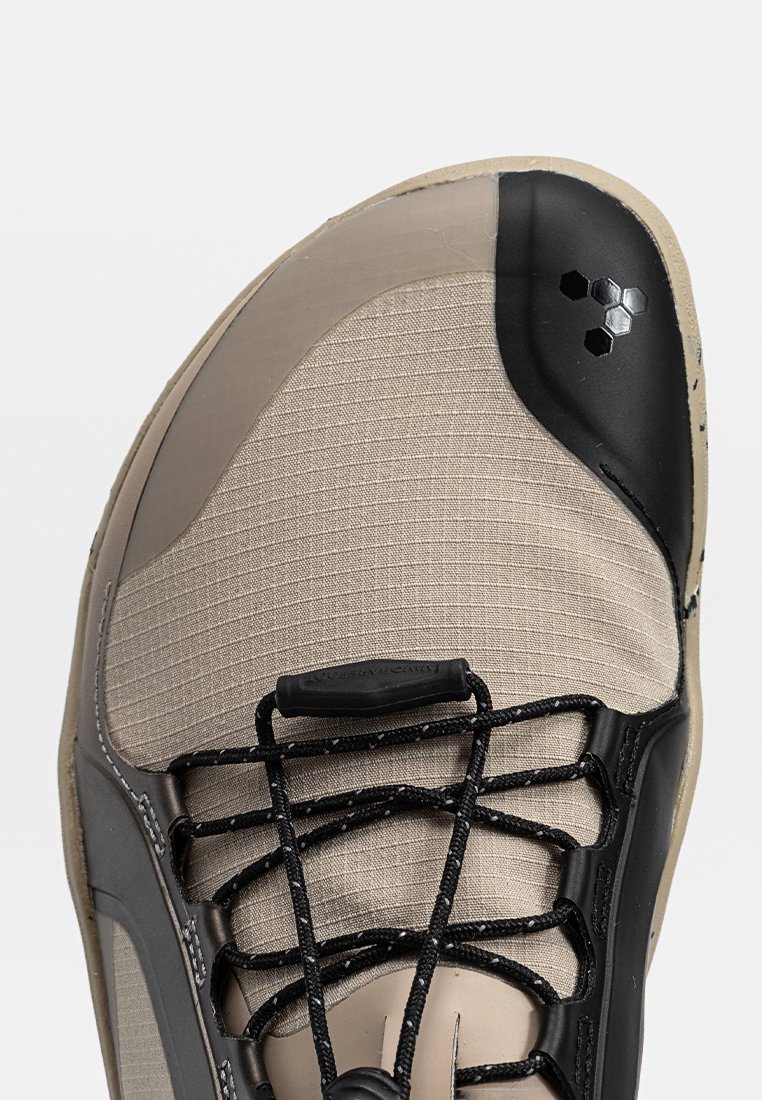Zapatilla deportiva beige de múltiples texturas con una punta negra, que presenta una forma redondeada, cordones negros y un detalle de logo hexagonal.