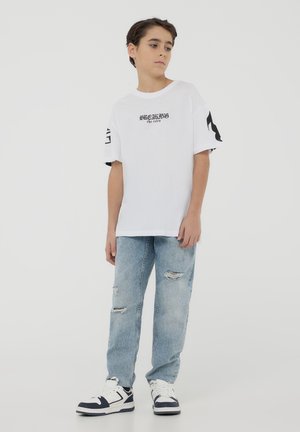 T-shirt bianco a maniche corte con grafiche testuali nere, abbinato a jeans leggeri azzurri strappati e sneakers bianche e nere.