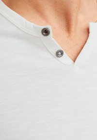 Weißes Kurzarm-Henley-Shirt mit V-Ausschnitt, ausgestattet mit zwei dekorativen grauen Knöpfen am Kragen, aus weichem Stoff mit leichter Textur.
