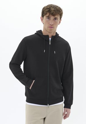 Zwarte zip-up hoodie van zacht materiaal, met een capuchon, voorzakken en geribbelde mouwen. Witte ondershirt en beige broek zichtbaar.