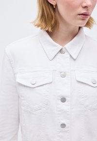 Κοντινό πλάνο ενός ατόμου που φοράει λευκό τζιν μπουφάν με ασημένια κουμπιά και χρυσούς κρίκους σκουλαρίκια, δείχνοντας το γιακά και την περιοχή του στήθους.