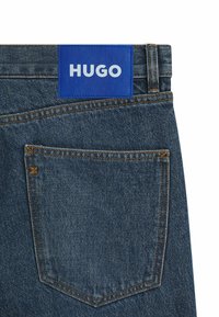 Denim jeans feature a blue leather waistband label with "HUGO" printed in white. The fabric is medium blue with orange stitching and a rear pocket.