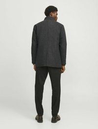 Un manteau en laine gris foncé avec un col haut, une seule couture arrière, associé à un pantalon noir et des chaussures brunes, présenté de dos.