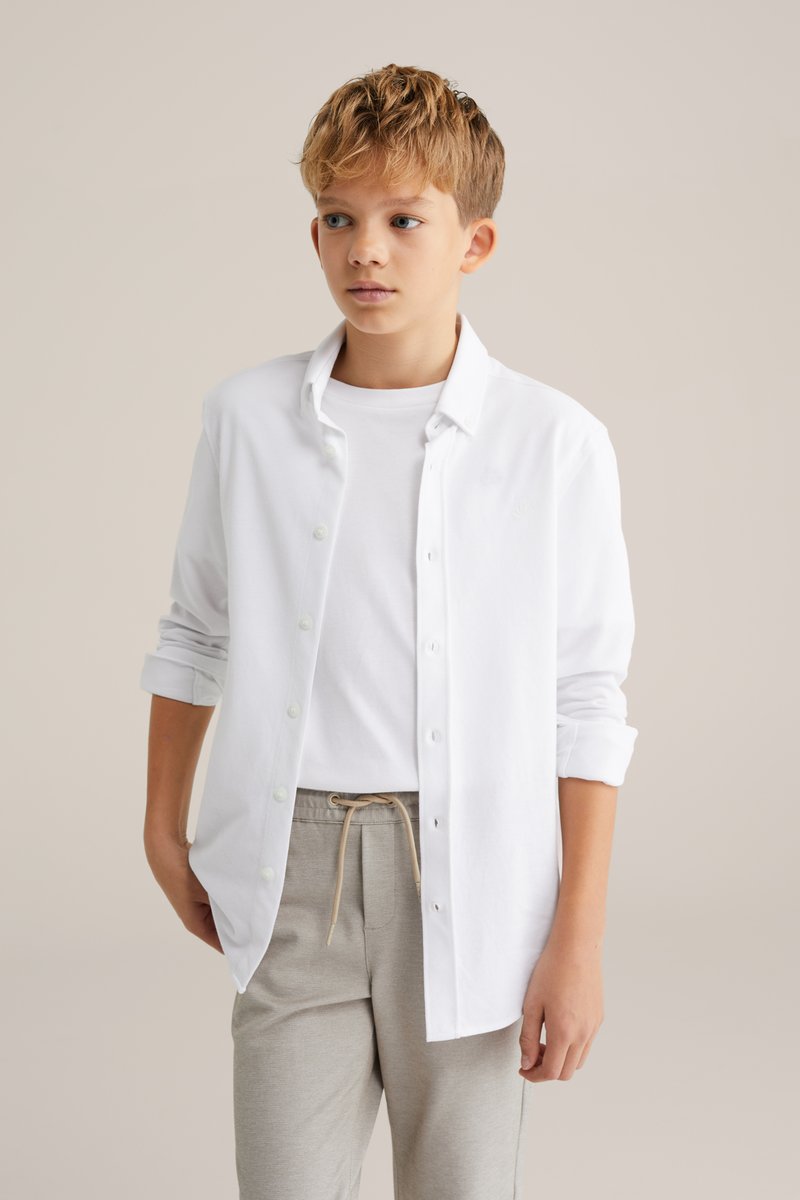 Witte blouse over een wit t-shirt, gedragen met lichtgrijze broek en een beige trekkoord. Stof lijkt zacht en licht van gewicht.