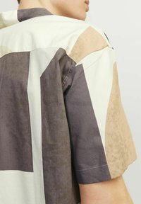 Cămașă cu mânecă scurtă dintr-un material moale, cu forme abstracte în nuanțe de bej stins, maro și crem; include cusături contrastante și un guler lejer.
