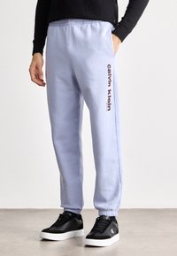 Ανοιχτό μπλε φόρμες με ελαστική μέση και μανσέτες, με ένα μπορντό λογότυπο "Calvin Klein" στο πλάι, συνδυασμένες με μαύρα αθλητικά παπούτσια.