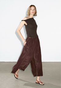 Top nero senza spalle abbinato a pantaloni larghi in satin bordeaux; modella che indossa sandali piatti rossi, design minimalista, texture liscia.