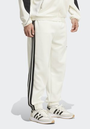 Muž oblečený v bílých sportovních kalhotách se černými pruhy po stranách a odpovídajících bílých a černých teniskách adidas, stojící na šedé podlaze.