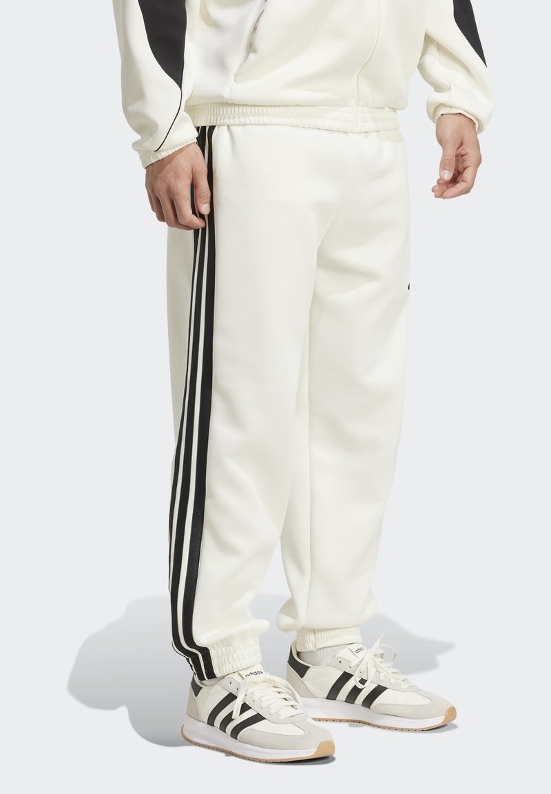 Muškarac u bijelim sportskim hlačama s crnim prugama sa strane i odgovarajućim bijelim i crnim adidas tenisicama, stoji na sivom podu.