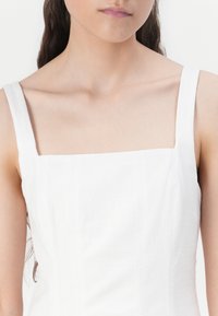 Robe blanche à encolure carrée avec des bretelles larges, un tissu texturé et un corsage ajusté. Design simple et minimaliste, adapté à diverses occasions.