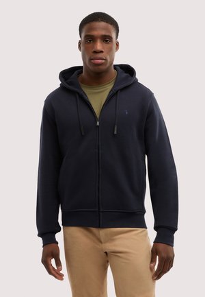 Junger Mann, der einen marineblauen Kapuzenpullover mit Reißverschluss über einem olivgrünen Hemd und beigen Hosen trägt, vor einem schlichten hellen Hintergrund stehend.