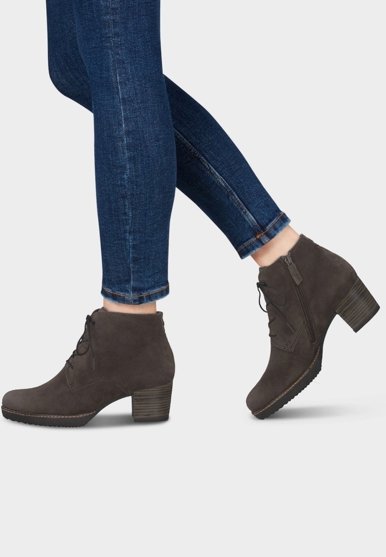 Personne portant des bottines en daim marron foncé à talons blocs et un jean skinny bleu, les orteils pointés vers le bas sur un pied.