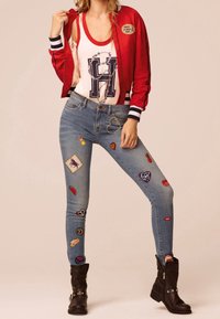 Röd bombarjacka med randiga mudd, vit linne med en stor bokstav, och blå jeans prydda med färgglada lappar samt mörkbruna stövlar.