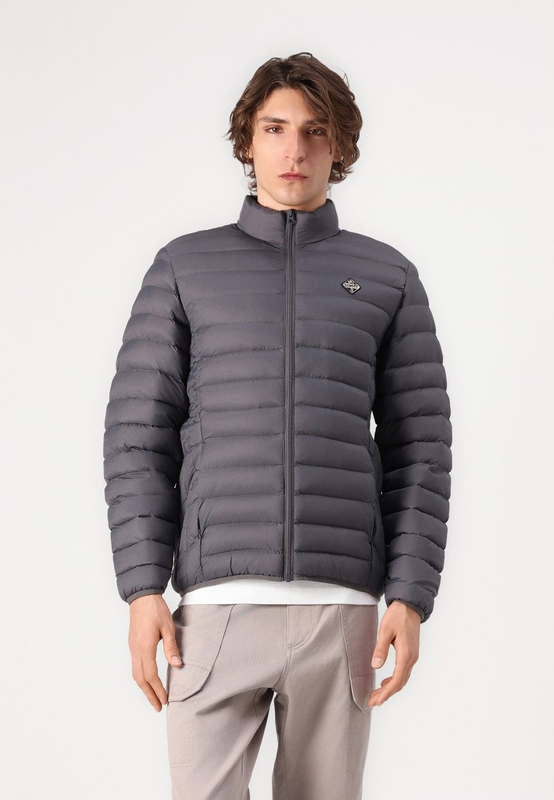 Manteau rembourré isolé gris avec un col haut, un design matelassé horizontal et un petit logo sur la poitrine. Le tissu a une texture lisse.