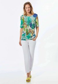 Kleurige, korte mouwen top met een bloemen- en abstracte print in groen, blauw en beige, gecombineerd met witte broek en gele sandalen.