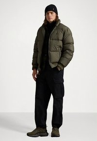 Alyvuogių žalios spalvos pūkinė striukė su aukštu apykakle, ribotomis rankogaliais ir dviem priekinėmis kišenėmis, derinama su juodomis krovinių kelnėmis ir tamsiais sportiniais bateliais.