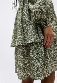 Κοντινό πλάνο ενός ατόμου που φορά ένα φόρεμα με πράσινο και άσπρο φλοράλ σχέδιο και μακριά μανίκια, με το χέρι να ακουμπά απαλά στο πλάι.