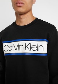 Czarna bluza z białym paskiem z napisem "Calvin Klein" w pogrubionych czarnych literach, ozdobiona dwoma niebieskimi paskami na wysokości klatki piersiowej.