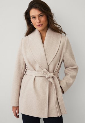 Vrouw draagt een beige wollen jas met riem en brede revers, staand tegen een effen lichte achtergrond, handen ontspannen langs haar zij.