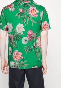 Camicia a maniche corte in tessuto verde brillante con un motivo floreale che presenta peonie rosa e bianche, foglie e una vestibilità comoda.