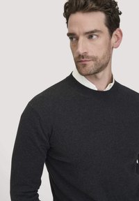Dunkelgrauer Strickpullover mit Rundhalsausschnitt und gerippten Bündchen, getragen über einem weißen Hemd mit Kragen. Glatte Textur, figurbetontes Design.