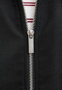 Primo piano di una cerniera argentata parzialmente aperta su tessuto nero, che svela una camicia bianca con righe orizzontali rosse sotto.