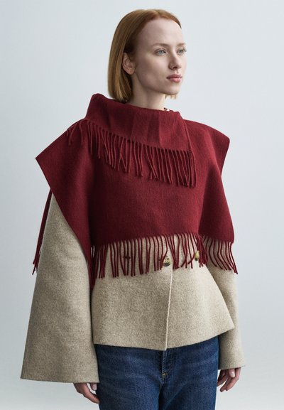 Vícevrstvá svrchní oděv sestávající z vínově červeného vlněného vrchního dílu s třásněmi a béžové vlněné spodní části, představující široké rukávy a knoflíkové detaily.