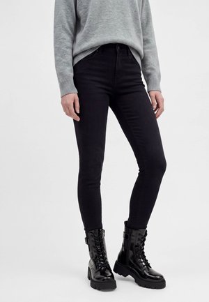 Personne portant un jean skinny noir taille haute, un pull gris et des bottes de combat noires brillantes à lacets, debout devant un fond clair uni.