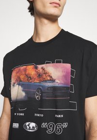 Svart bomullströja med en stor grafisk bild av en blå bil med eld och rök. Texten nedanför inkluderar stadsnamn och en jordglobikon.