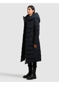 Cappotto imbottito nero con design a zig zag, dotato di un ampio cappuccio, chiusura con zip e tasche laterali. Indossato sopra leggings neri e stivali con tacco.