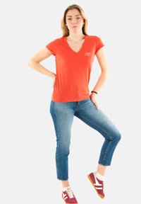 T-shirt rouge à col en V et à manches courtes avec un petit logo, associé à un jean skinny bleu moyen et des baskets bordeaux avec des accents blancs.