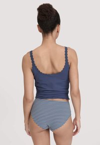 Vrouw met krullend haar in een buns, gekleed in een marineblauwe top met kanten schouderbanden en blauw-grijze gestreepte hoge tailleonderbroeken, staande met de rug naar de camera.