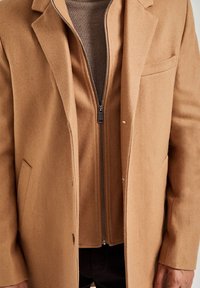 Kamelull-blandning kappa med skalkragar, sidofickor och ett synligt brunt dragkedjejinnerlager med en texturerad tyg under.