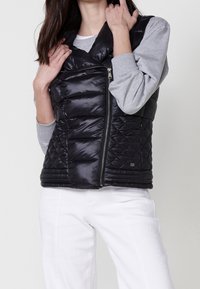 Femme portant un gilet matelassé sans manches noir par-dessus une chemise grise à manches longues et un pantalon blanc, tenant le col du gilet avec les deux mains.