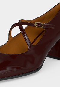 Chaussure Mary Jane en cuir verni rouge foncé avec un petit talon bloc et une bride à boucle dorée sur le dessus.