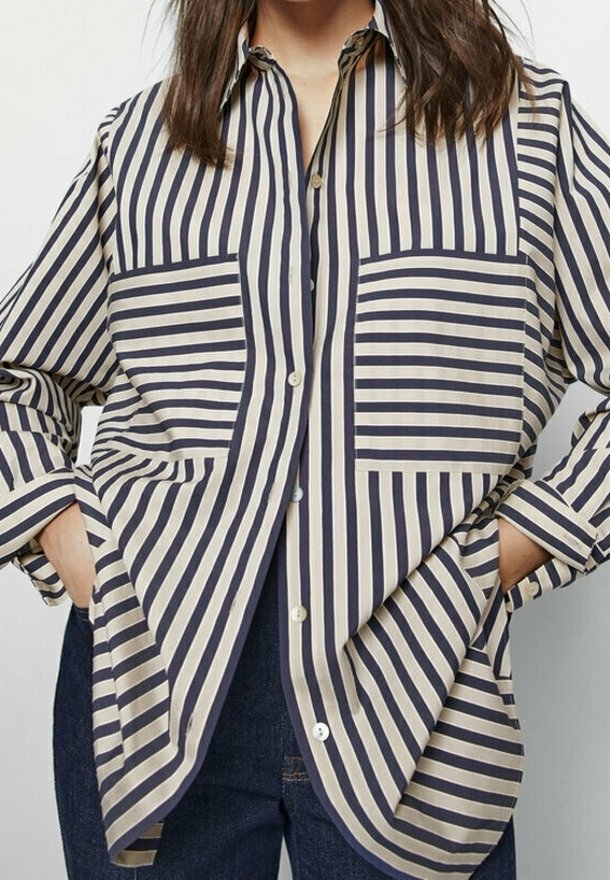 Femme portant une chemise ample à manches longues à rayures verticales bleu marine et beige avec des poches plaquées, mains dans les poches.