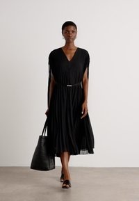 Black pleated dress with a V-neck and cape sleeves, cinched at the waist with a silver belt. Model holds a black tote bag. Smooth texture.