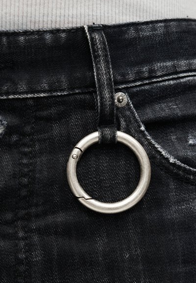 Ασημένιο μεταλλικό κρίκο κλειδιών προσαρτημένο σε μαύρο λουράκι τζιν πάνω από την μπροστινή τσέπη μαύρων τζιν με λευκά ραφές.
