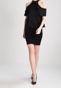 Top negro sin mangas con volantes, combinado con una falda corta negra ajustada. Tejido suave y silueta elegante, complementado con tacones negros.