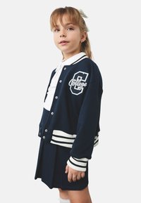 Marine varsityjack met witte ribgebreide accenten en geborduurd "C"-logo, gedragen over een wit overhemd met kraag en een donkere geplooide rok.