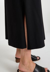 Jupe en coton noir avec une fente latérale, présentant un ourlet propre. Associée à des sandales noires à glissière avec une bride texturée et un talon en bois.