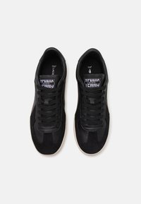 Zapatillas negras de cuero y ante, con un acabado texturizado, puntera redondeada y cordones planos. Etiqueta de la marca "TENNIS LACOSTE" visible.