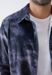Camisa de pana en azul oscuro con patrones de tie-dye. Presenta un cierre frontal de botones y un cuello clásico. Textura suave con diseño acanalado.