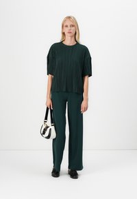 Tamna zelena plisirana bluzica sa širokim rukavima, uparena s odgovarajućim širokim hlačama. Crne cipele i mala crno-bijela torbica upotpunjuju ovaj outfit.